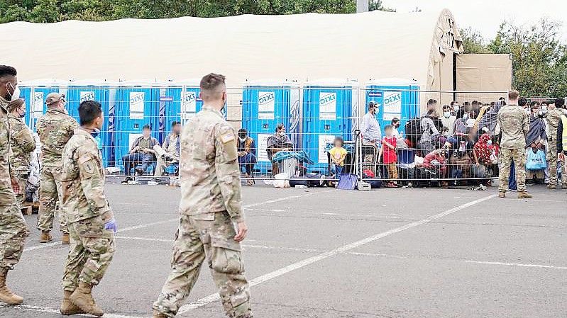 Aus Afghanistan evakuierte Menschen stehen auf der Ramstein Air Base vor einem Zelt. Davor gehen US-Soldaten. Foto: Uwe Anspach/dpa