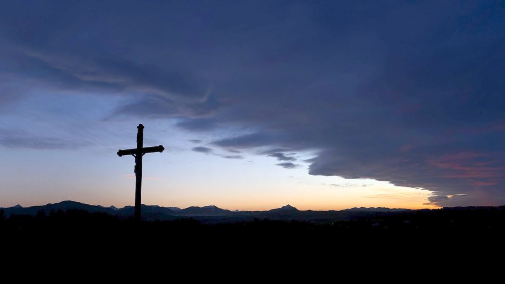 Eine Wolkenfront zieht hinter einem Wegkreuz über die im Sonnenuntergang liegende Berglandschaft. Foto: dpa/Karl-Josef Hildenbrand