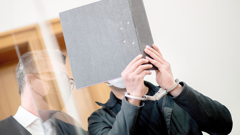 Der Angeklagte steht zu Prozessbeginn mit Handfesseln in einem Saal im Landgericht und bedeckt sein Gesicht mit einem Aktenordner. Foto: Dittrich/dpa
