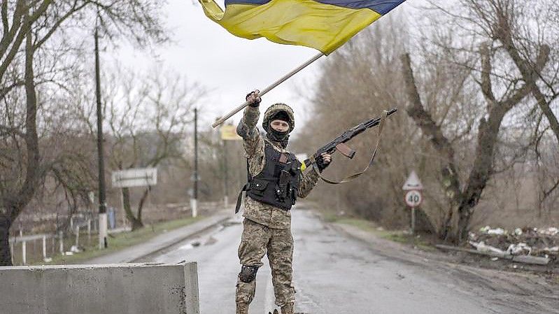 Ein ukrainischer Soldat steht an einer Straßenblockade und hält die ukrainische Flagge hoch. Foto: Rodrigo Abd/AP/dpa