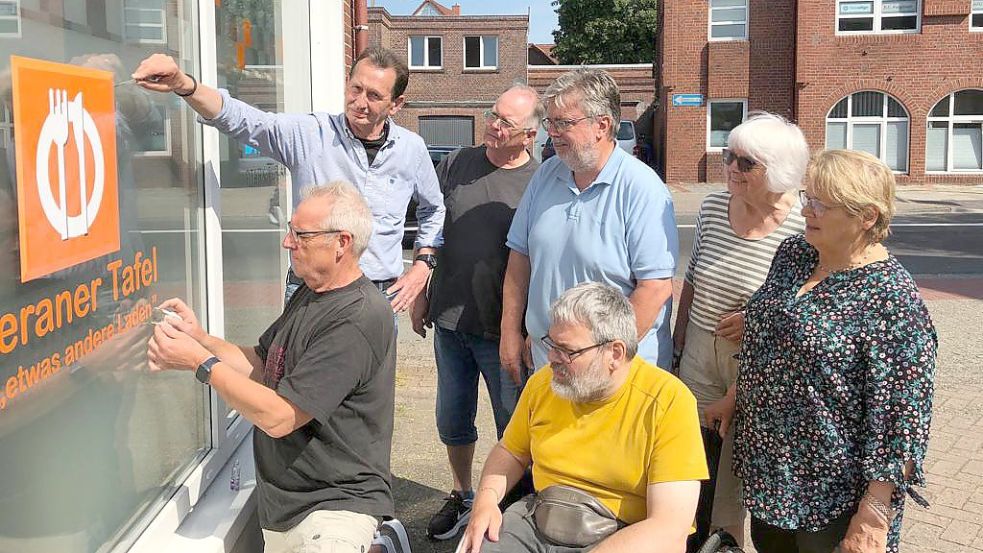 Noch tun die Helfer der Leeraner Tafel nur so, als ob sie das Logo der Tafel von der Scheibe ihres jetzigen Domizils abkratzen. In wenigen Monaten werden Hanna Weidner (von rechts), Heidi Lünemann, Andreas Poppen (vorne), Andreas Bartels, Wolfgang Mallik, Johannes Pauw (hinten) und Detlev Hartmann tatsächlich Hand anlegen. Foto: Mielcarek