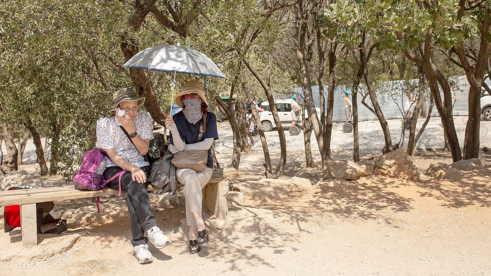 Touristen sitzen unter Bäumen am Eingang der Akropolis in Athen. Foto: Socrates Baltagiannis/dpa