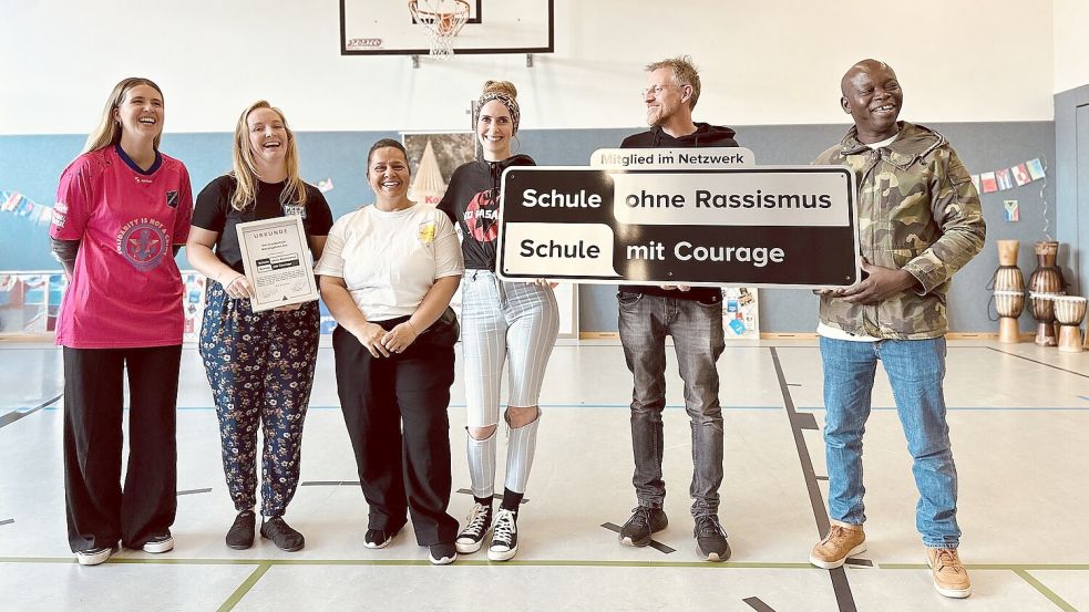 Mit Schild und Urkunde präsentieren sich (von links): Sina Cordes (Projektleiterin an der Grundschule), Sophie Eickhorst (Netzwerk „Schule ohne Rassismus, Schule mit Courage“), Bilmez Kaya (Yezidischer Kulturverein), Annabel Kone (Patin des Projekts), Schulleiter Michael Markus und Ali Kone (Afrikanische Diaspora Ostfriesland und Pate des Projekts). Foto: privat