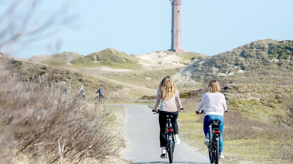Zwei junge Frau fahren mit Elektrorädern eines Fahrradverleihs auf einem Weg durch die Dünen der Insel. Im Hintergrund ist der Leuchtturm von Norderney zu erkennen. Foto: Hauke-Christian Dittrich/dpa