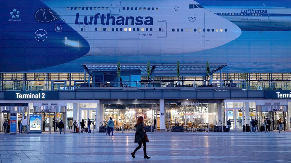 Passagiere gehen am Münchner Flughafen vor dem Terminal 2 an einem Plakat mit Flugzeugen der Lufthansa entlang. Foto: Sven Hoppe